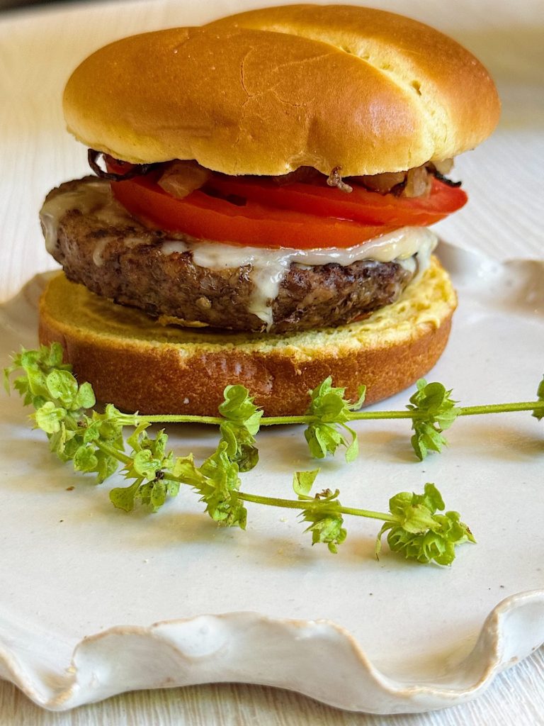 A cheeseburger with tomato slices, melted cheese, and grilled onions on a toasted bun sits on a white plate beside a sprig of fresh green herbs—perfect for easy fall dinners.