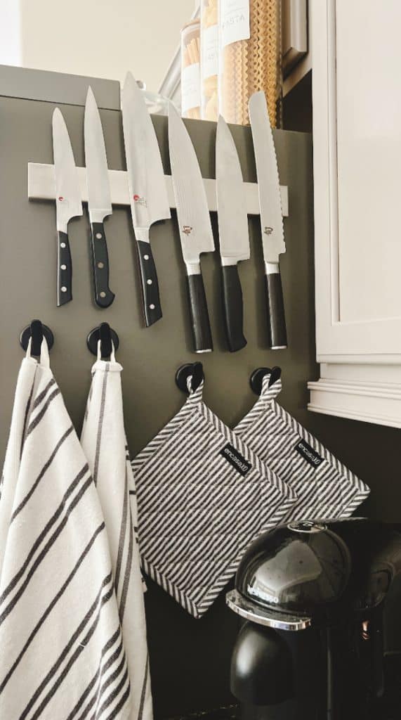 A magnetic strip holds five kitchen knives above two striped dish towels and pot holders hanging on hooks—perfect for prepping easy fall dinners; pasta jars line the shelf above, with a coffee maker on the counter below.