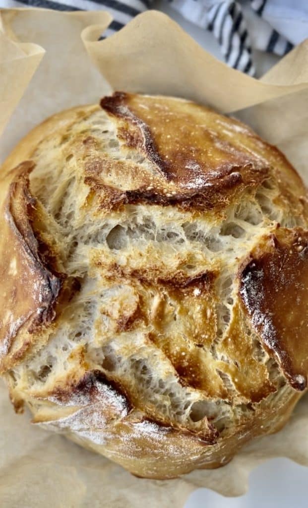 A round loaf of rustic, golden-brown artisan bread with a crisp, cracked crust sits on parchment paper, showing its airy interior—perfect for pairing with easy fall dinners. A striped kitchen towel is partially visible in the background.