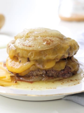 A stack of cheesy, melted French onion soup burgers inspired by the In-N-Out Burger Recipe (Flying Dutchman), topped with caramelized onions and gooey cheese, with savory juices and melty cheese pooling at the bottom.