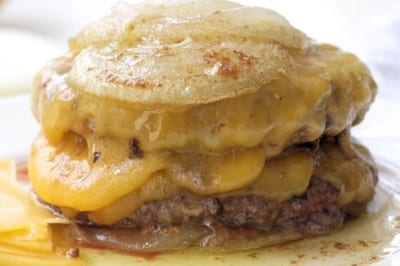 A stack of cheesy, melted French onion soup burgers inspired by the In-N-Out Burger Recipe (Flying Dutchman), topped with caramelized onions and gooey cheese, with savory juices and melty cheese pooling at the bottom.