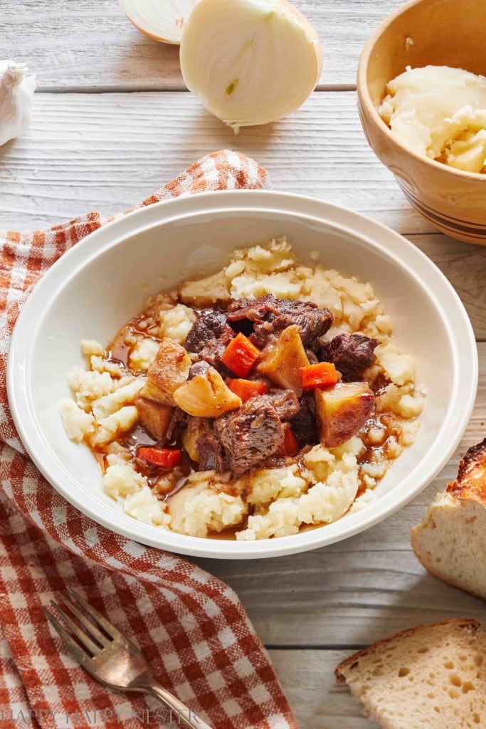 A bowl of mashed potatoes topped with beef stew, filled with carrots and potatoes, sits on a checkered napkin—perfect for easy fall dinners. Nearby, sliced bread, an onion, and extra mashed potatoes rest on a rustic wooden table.