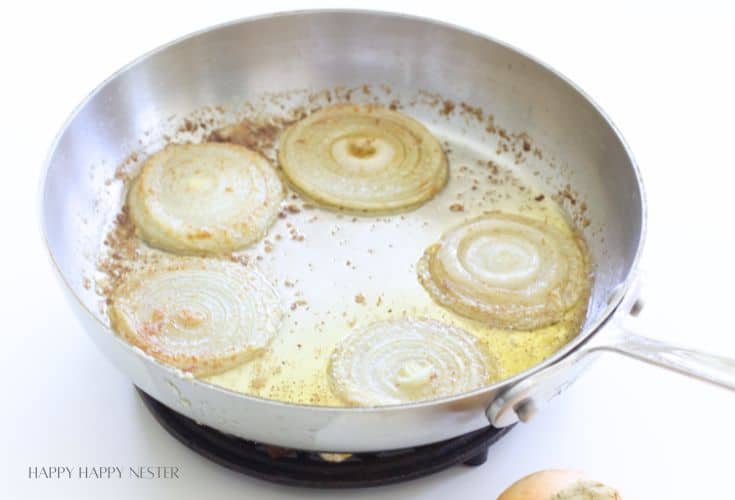 Five slices of onion are being sautéed in a stainless steel pan with oil, developing light browning—perfectly prepped. The pan rests on a white surface.
