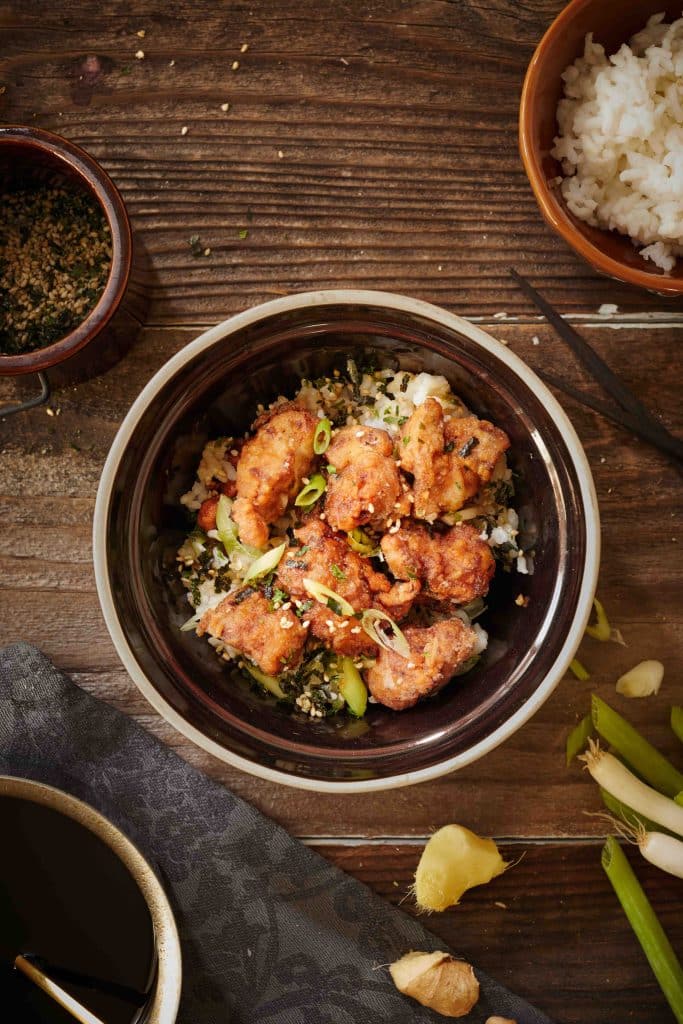 A bowl of crispy fried chicken pieces served over rice, garnished with chopped green onions—perfect for easy fall dinners. Nearby are a bowl of white rice, seasoning, green onions, and garlic on a wooden table.