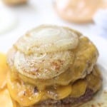 A close-up of a cheeseburger inspired by the In-N-Out Burger Recipe (Flying Dutchman), with two beef patties, melted cheddar cheese, and thick slices of cooked onion on top, cheese slices and condiments blurred in the background.