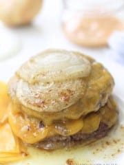 A close-up of a cheeseburger inspired by the In-N-Out Burger Recipe (Flying Dutchman), with two beef patties, melted cheddar cheese, and thick slices of cooked onion on top, cheese slices and condiments blurred in the background.