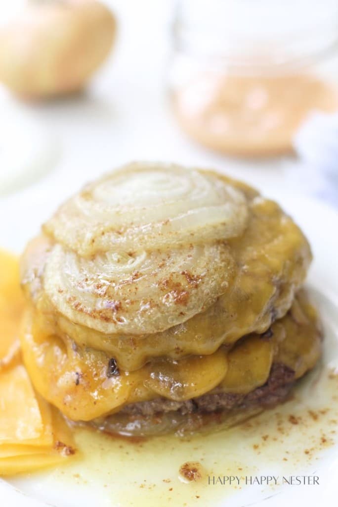 A close-up of a cheeseburger with two beef patties, melted cheddar cheese, and thick slices of cooked onion on top, cheese slices and condiments blurred in the background.