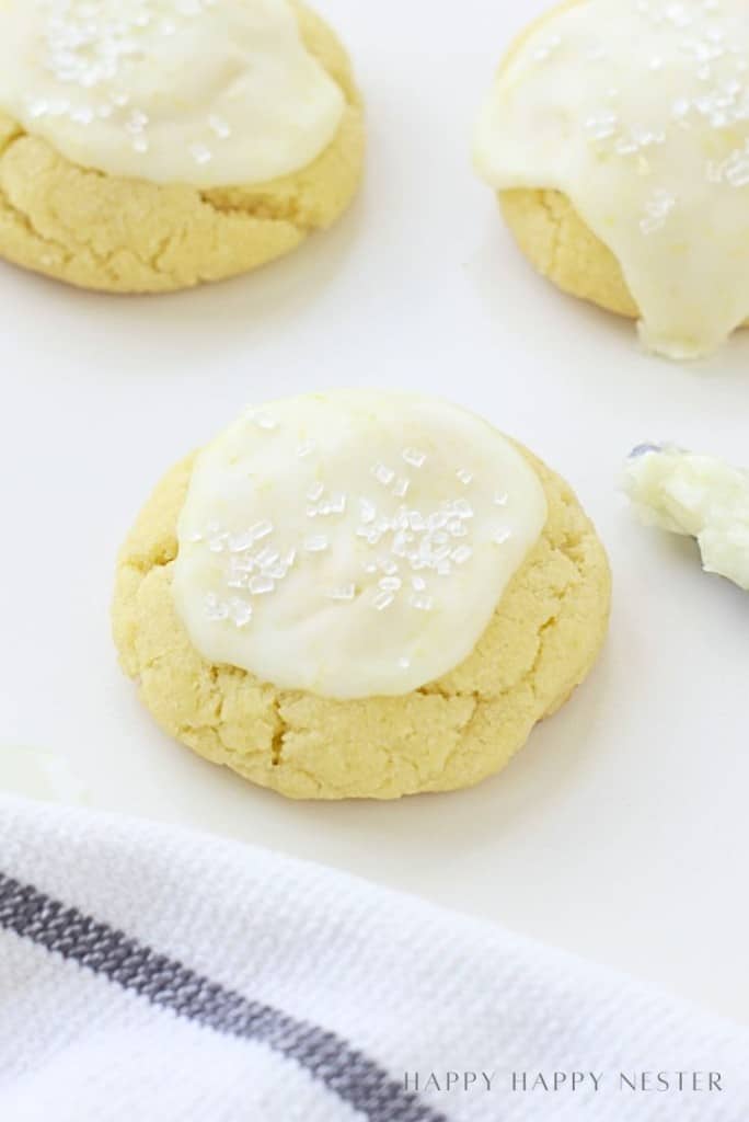 A soft, round lemon sugar cookie recipe topped with white icing and coarse sugar crystals, resting on a white surface with part of another cookie and a striped cloth nearby.