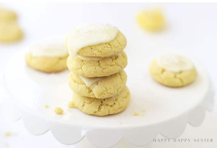 A stack of four yellow cookies with white icing on top sits on a white cake stand. Other cookies, also topped with icing, are blurred in the background. The scene is bright and minimalistic.