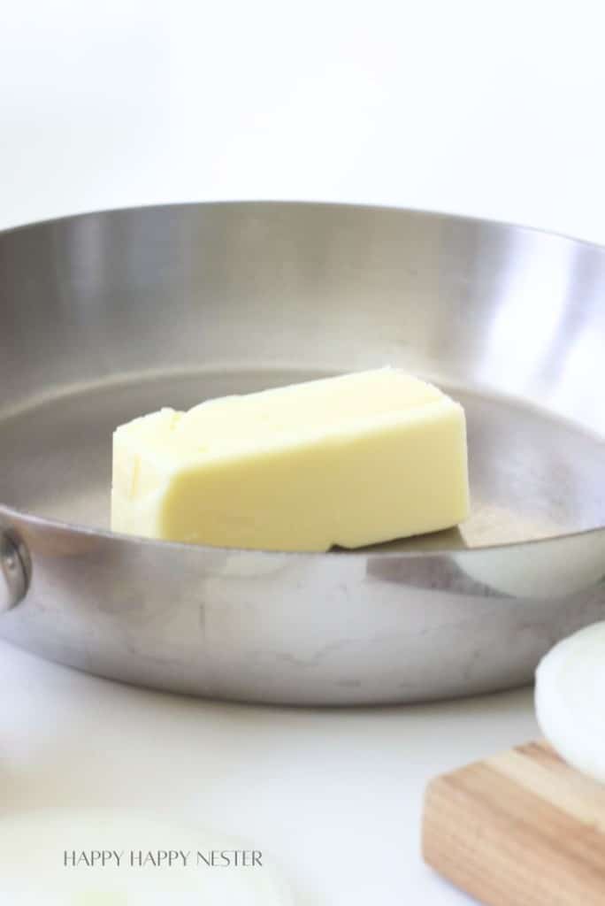 A stick of butter sits in a stainless steel pan with a white background, evoking the start of an In-N-Out Burger Recipe (Flying Dutchman). A partial cutting board and an onion are visible in the corner.