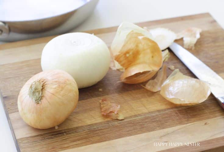 Two onions, one peeled and one unpeeled, sit on a wooden cutting board next to a kitchen knife—perfect prep for an In-N-Out Burger Recipe (Flying Dutchman). A metal pan is partially visible in the background.