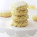 A stack of four soft, yellow cookies made with our lemon sugar cookie recipe sits on a white cake stand, topped with white icing and sprinkles, with more cookies blurred in the background.