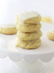 A stack of four soft, yellow cookies made with our lemon sugar cookie recipe sits on a white cake stand, topped with white icing and sprinkles, with more cookies blurred in the background.