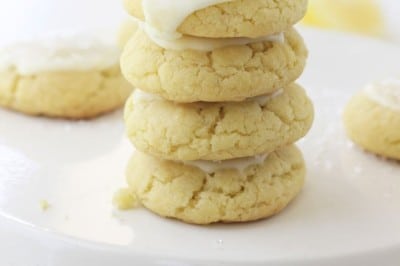 A stack of four soft, yellow cookies made with our lemon sugar cookie recipe sits on a white cake stand, topped with white icing and sprinkles, with more cookies blurred in the background.