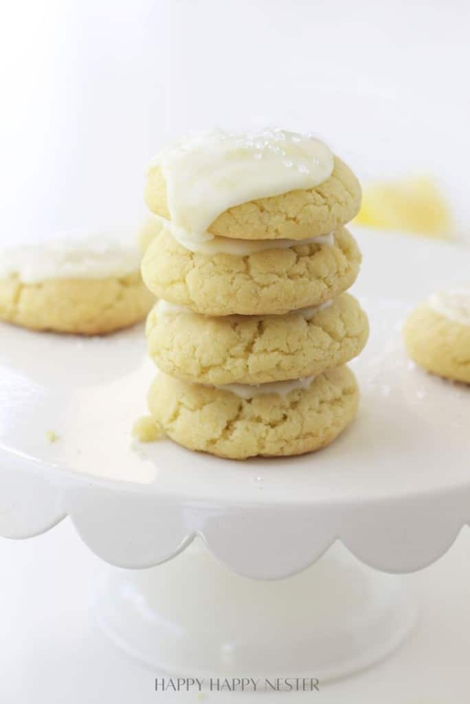A stack of four soft, yellow cookies made with our lemon sugar cookie recipe sits on a white cake stand, topped with white icing and sprinkles, with more cookies blurred in the background.