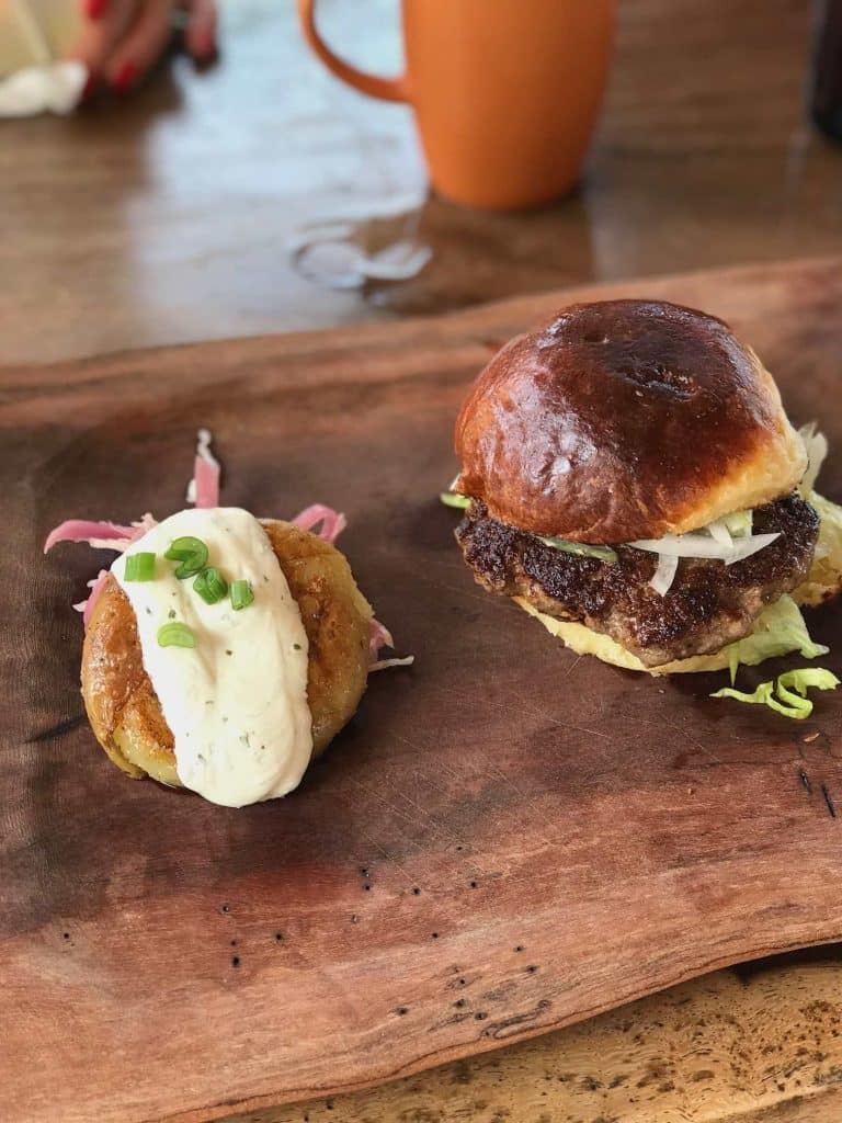 A burger with lettuce and cheese on a glossy bun sits on a wooden board next to a golden fried patty, reminiscent of the flying dutchman burger recipe, topped with creamy sauce and chopped green onions. A hand and an orange mug are blurred in the background.