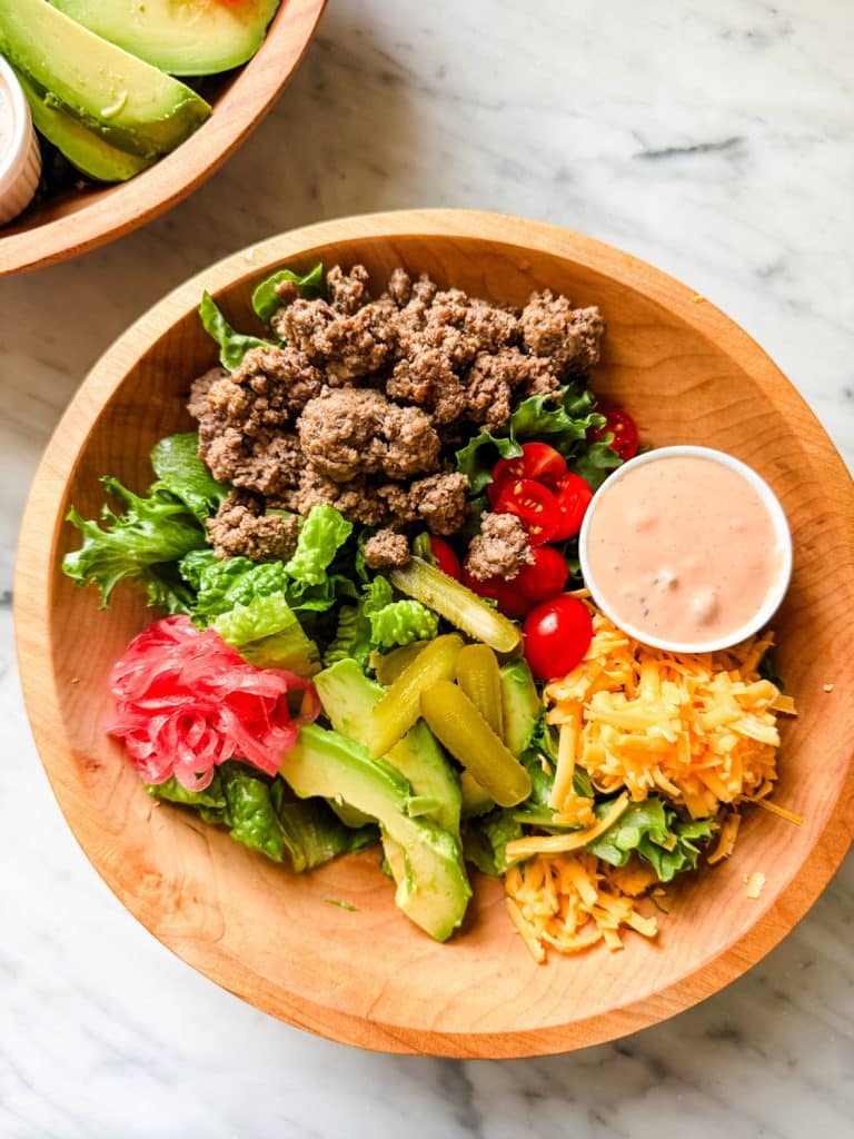 A wooden bowl filled with ground beef, mixed greens, cherry tomatoes, shredded cheddar cheese, sliced pickles, avocado, pickled onions, and a cup of creamy dressing—perfect for those inspired by the flying dutchman burger recipe.