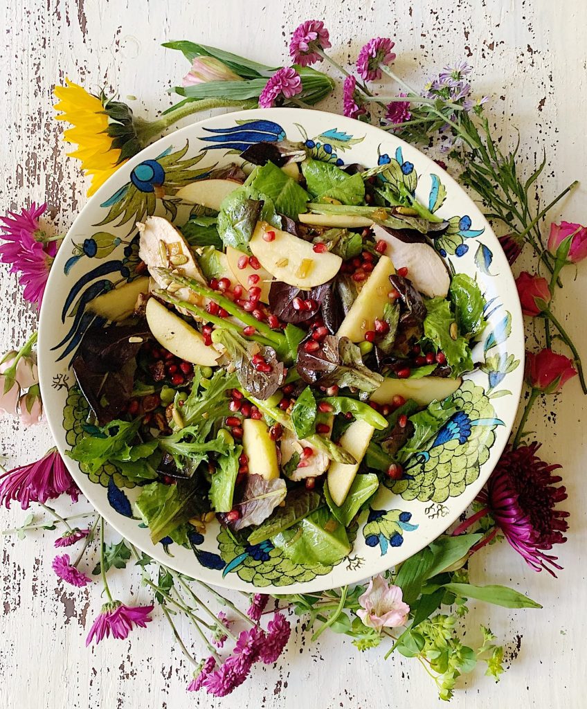 A colorful salad with mixed greens, apple slices, and pomegranate seeds in a decorative bowl—one of my favorite Thanksgiving recipes—surrounded by scattered fresh flowers on a rustic white wooden surface.
