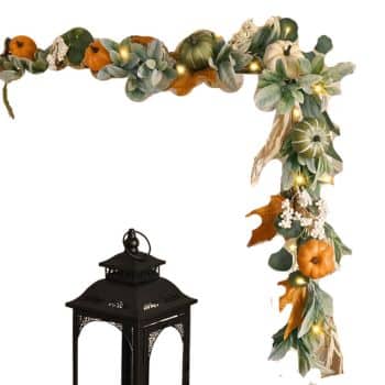 A decorative autumn garland with green leaves, small orange and green pumpkins, white berries, and string lights is draped over a corner, with a black metal lantern positioned beneath it.