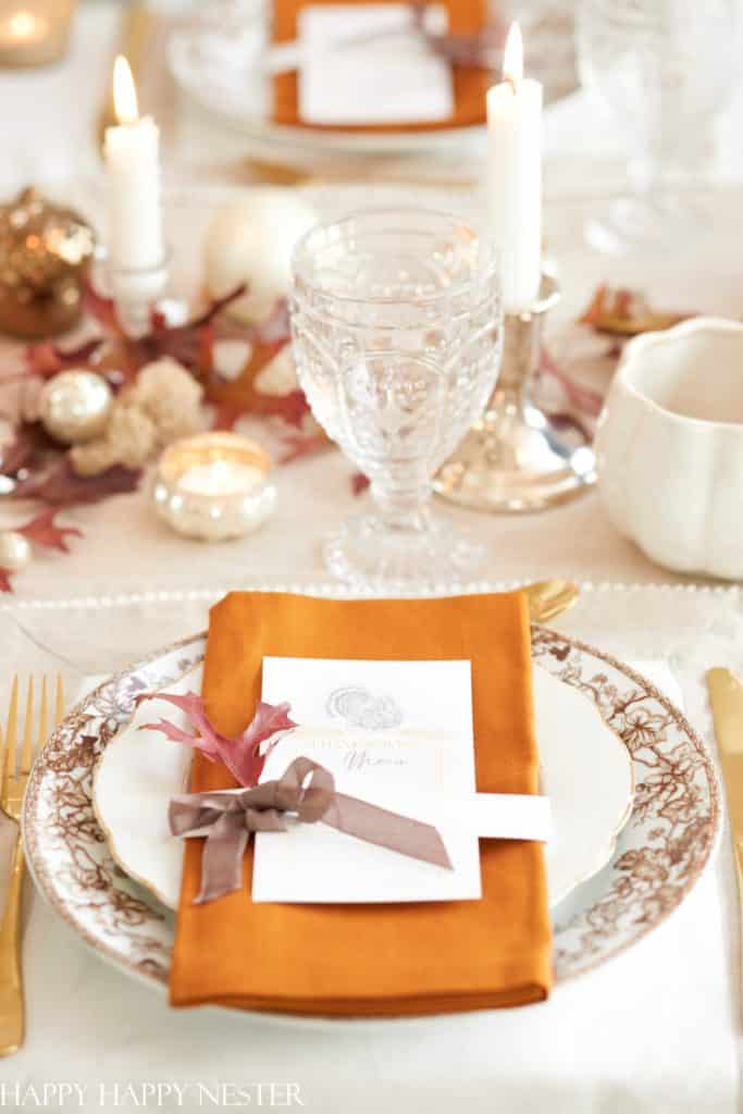 Elegant fall-themed table setting with an ornate plate, gold flatware, orange napkin, menu card featuring my favorite Thanksgiving recipes, and a brown ribbon. Decorated with autumn leaves, candles, and glassware on a white tablecloth.