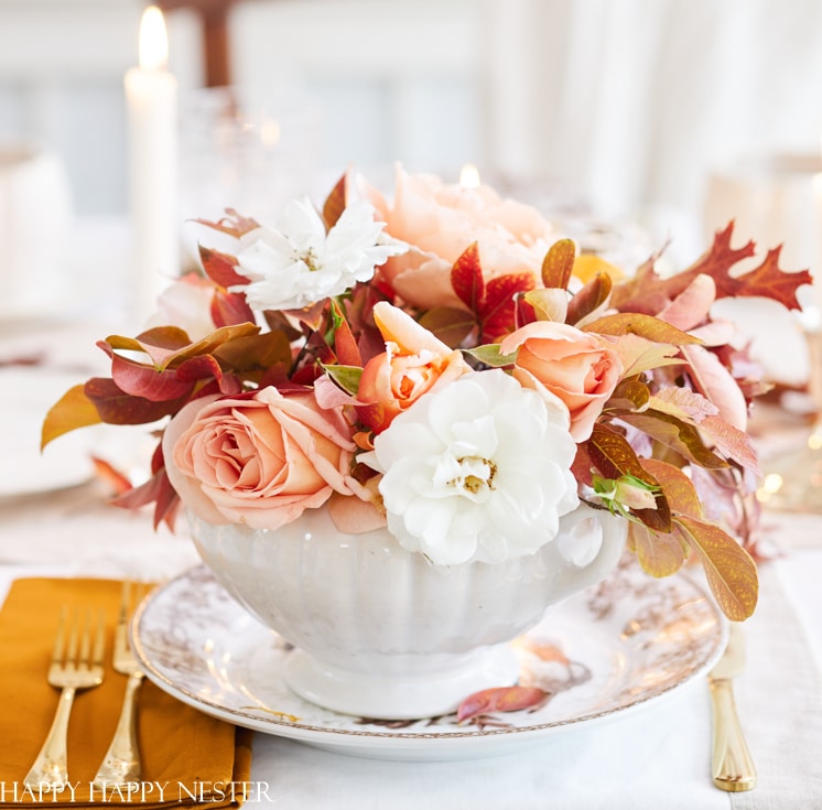 A white ceramic bowl filled with peach roses, white flowers, and autumn leaves sits on a patterned plate beside a gold fork and knife—the perfect setting for sharing my favorite Thanksgiving recipes, with candlelight softly blurred in the background.