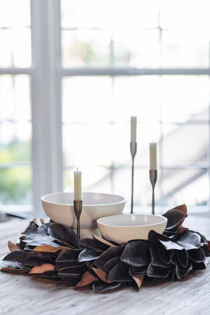 A decorative centerpiece features a Dough Bowl Candle, two white bowls, three tall white candles in metal holders, and a wreath of dark brown leaves, all arranged on a wooden table in front of a bright window.