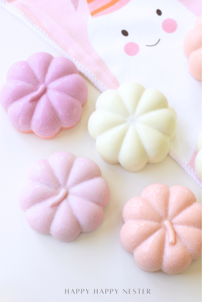 Three pastel-colored, pumpkin-shaped Japanese candies are arranged on a white surface, with a white and pink cloth featuring a cute face in the background—perfect inspiration for your next Chocolate Pumpkins or Halloween Recipe.
