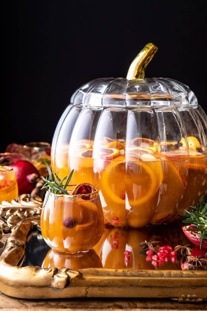 A pumpkin-shaped glass pitcher filled with orange punch, citrus slices, and pomegranate seeds sits on a gold tray. A matching glass garnished with rosemary and fruit is beside it—one of my favorite Thanksgiving recipes to serve guests.