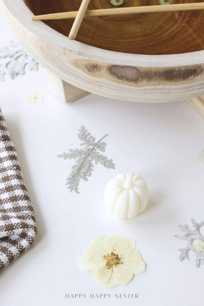 A small white decorative pumpkin, pressed flowers, and a brown wooden dough bowl candle diy are arranged on a white surface with a checkered cloth nearby. The words "HAPPY HAPPY NESTER" appear at the bottom.