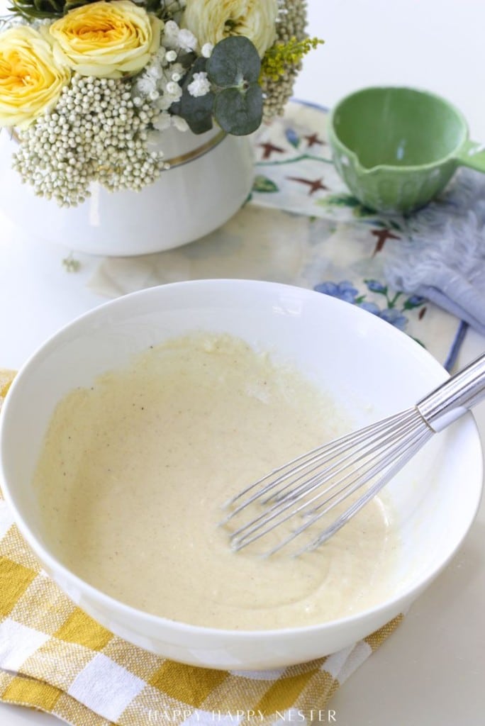 A metal whisk rests in a white bowl of batter on a yellow checkered cloth, with a floral arrangement and a green juicer in the background.
