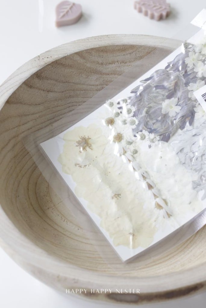 A wooden bowl holds a clear plastic-wrapped sheet with pressed white and purple flowers on it. Out of focus, pink leaf-shaped items are visible on the white surface nearby.