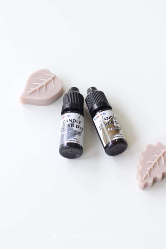Two bottles of candle liquid dye, one labeled "Black Grey" and the other "Brown," are displayed on a white surface next to two beige, leaf-shaped objects—perfect for your next dough bowl candle DIY project.