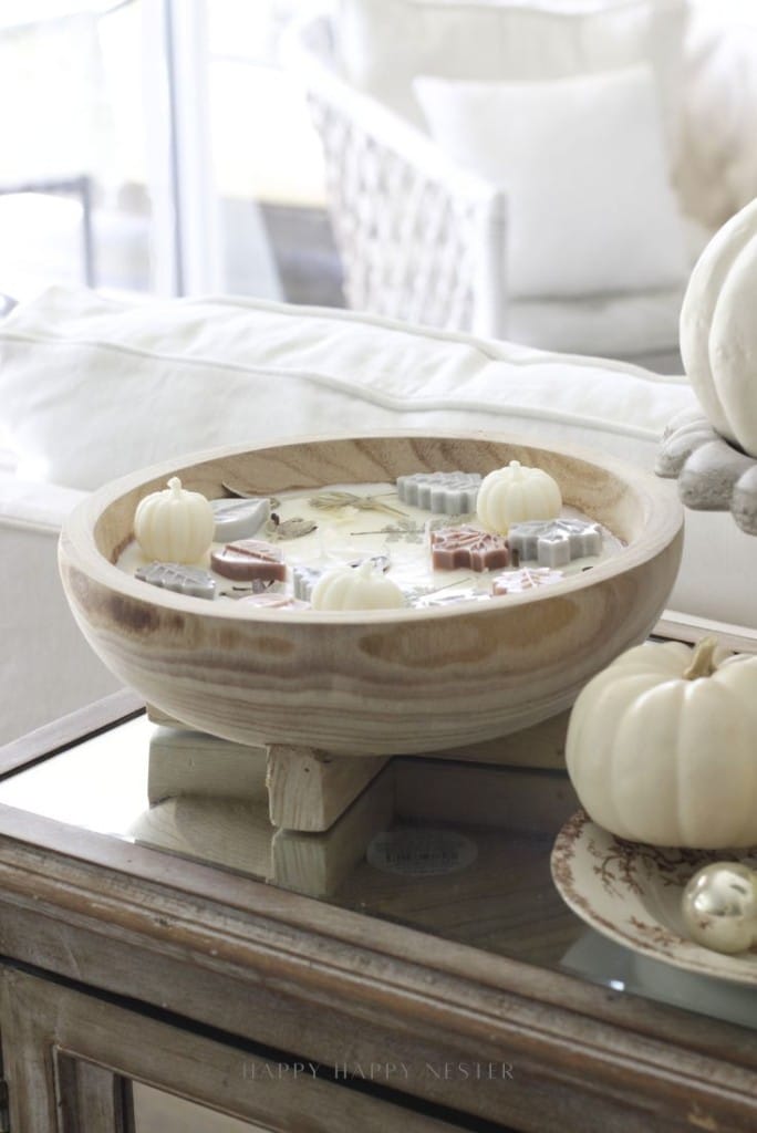 A large wooden bowl candle topped with small wax decorations shaped like pumpkins and leaves sits on a rustic wooden table, surrounded by white pumpkins and neutral cozy decor.