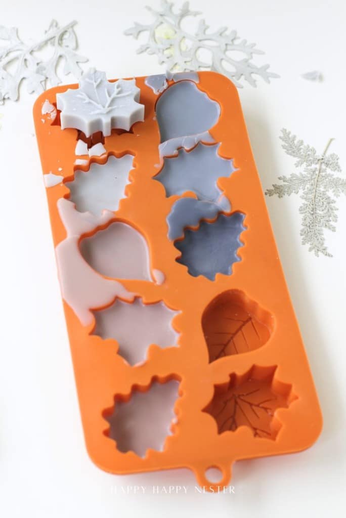 An orange silicone mold with various leaf-shaped cavities, some filled with gray and purple wax, sits on a white surface decorated with silver snowflake ornaments.