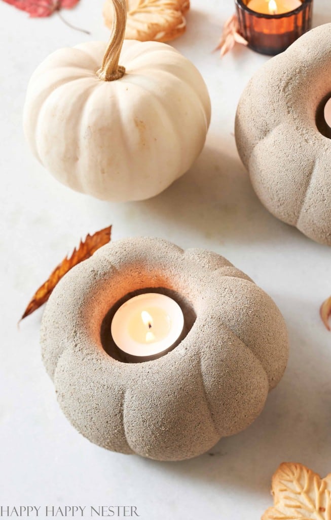 A lit tealight candle sits inside a textured, pumpkin-shaped candle holder next to a small white pumpkin and autumn leaves on a light surface—a charming scene for lovers of all things pumpkin.