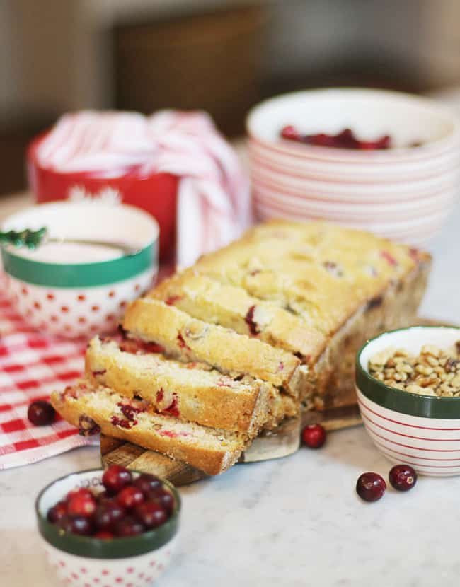 A loaf of cranberry bread, partially sliced, sits on a marble countertop surrounded by bowls of cranberries, nuts, and a festive mug—one of my favorite Thanksgiving recipes—set against a red-and-white checked cloth in the background.