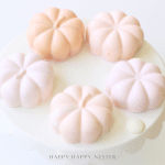 Five pastel pumpkin-shaped desserts sit on a round white cake stand, arranged in a circle. These chocolate pumpkins, in soft shades of pink and peach, create a delicate and elegant Halloween recipe presentation.