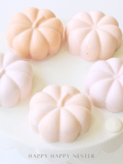 Five pastel pumpkin-shaped desserts sit on a round white cake stand, arranged in a circle. These chocolate pumpkins, in soft shades of pink and peach, create a delicate and elegant Halloween recipe presentation.