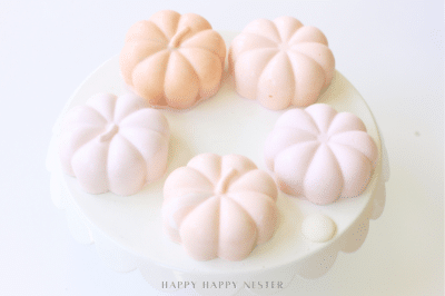 Five pastel pumpkin-shaped desserts sit on a round white cake stand, arranged in a circle. These chocolate pumpkins, in soft shades of pink and peach, create a delicate and elegant Halloween recipe presentation.