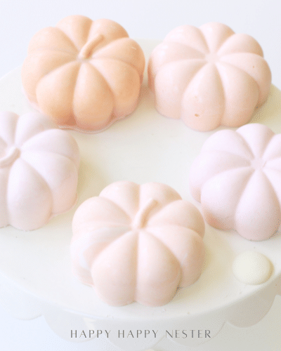 Five pastel pumpkin-shaped desserts sit on a round white cake stand, arranged in a circle. These chocolate pumpkins, in soft shades of pink and peach, create a delicate and elegant Halloween recipe presentation.