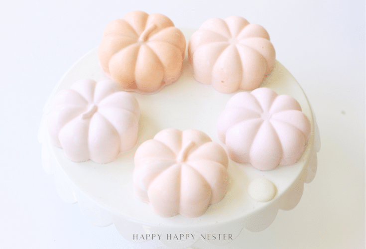 Five pastel pumpkin-shaped desserts sit on a round white cake stand, arranged in a circle. These chocolate pumpkins, in soft shades of pink and peach, create a delicate and elegant Halloween recipe presentation.