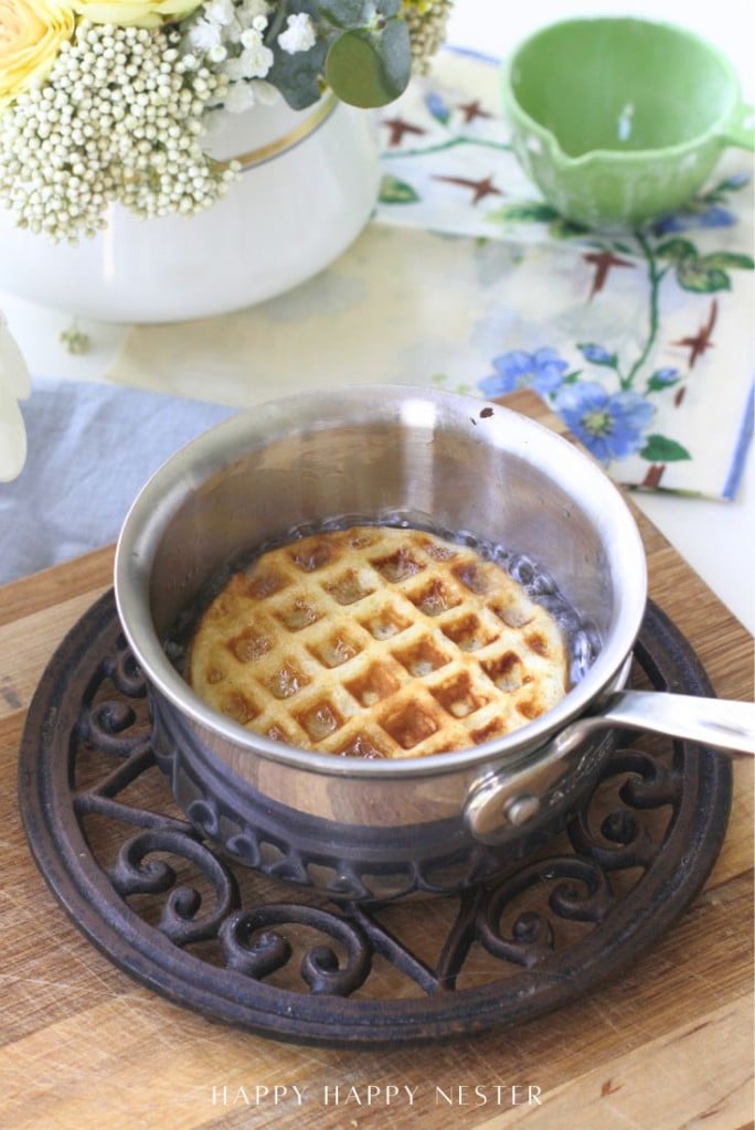A small waffle from a mini waffle donuts recipe is being cooked in a metal saucepan, placed on a round decorative trivet on a wooden board, with flowers and a green bowl in the background.