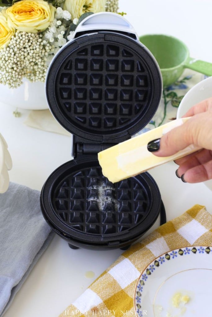 A hand holds a stick of butter, rubbing it onto an open, empty waffle maker. Flowers, a green cup, and a yellow-checkered napkin are visible on the table in the background.