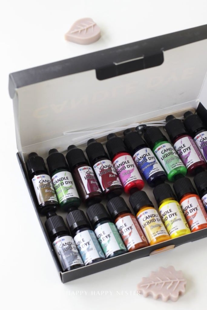 An open box displays bottles of candle liquid dye in various colors, each labeled with color names. Two pinkish wax leaf decorations are placed nearby on a white surface.