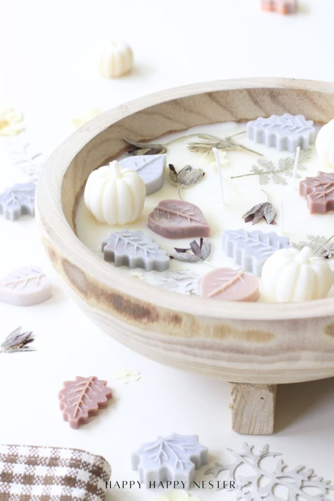 A rustic wooden bowl holds a white candle topped with wax decorations shaped like pastel leaves and pumpkins. Dried flowers and more wax leaf shapes are scattered around on a white surface.