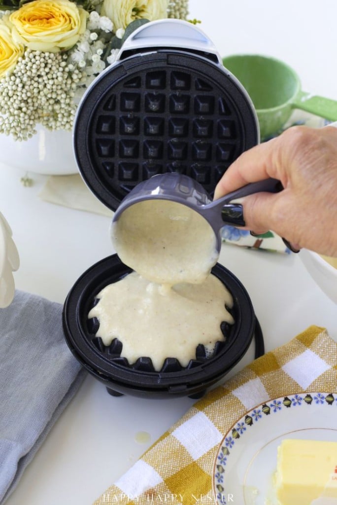 A hand pours waffle batter from a measuring cup into a black waffle maker on a white table, with flowers, a green mug, and a butter dish nearby.
