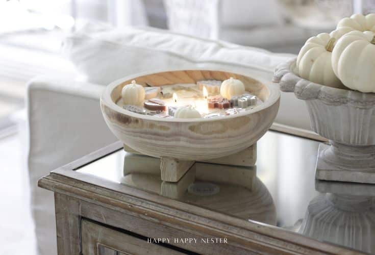 A handmade dough bowl candle with embedded mini pumpkins and autumn decorations sits on a rustic side table beside a ceramic bowl of white pumpkins in a cozy, bright living room.