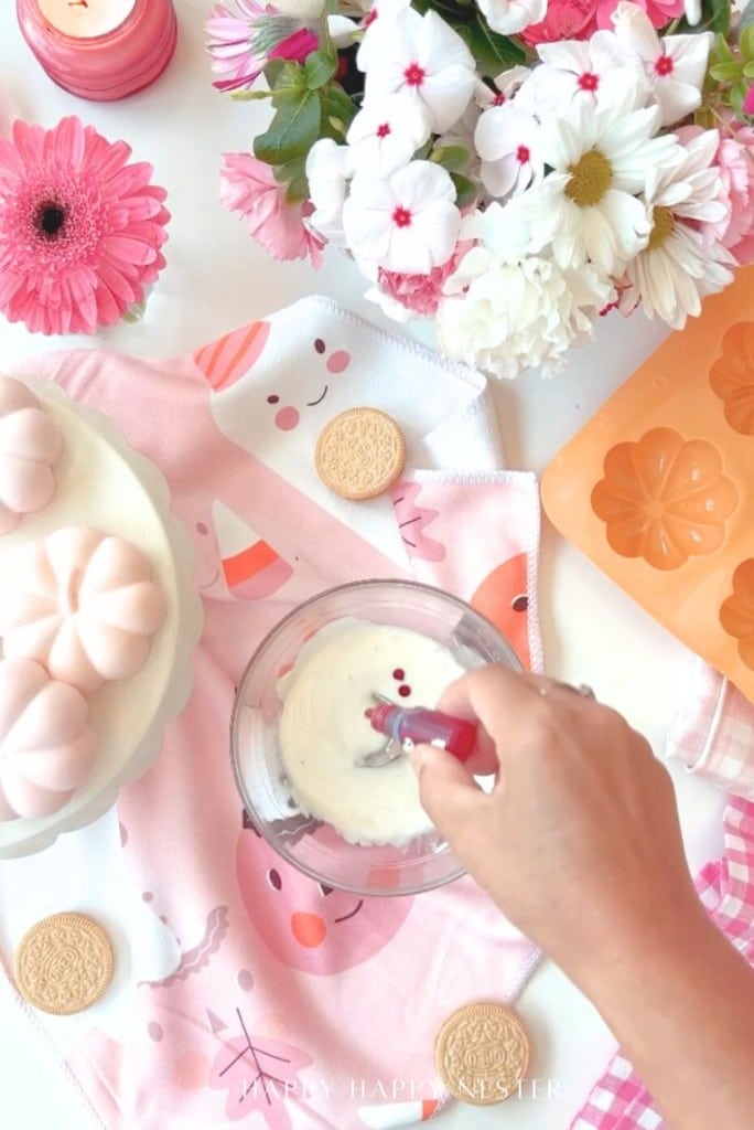 A hand mixes red food coloring into a bowl of white icing on a pink-themed table with flowers, cookies, a cake, a candle, and a pink napkin with cute faces. An orange flower-shaped silicone mold is also visible.