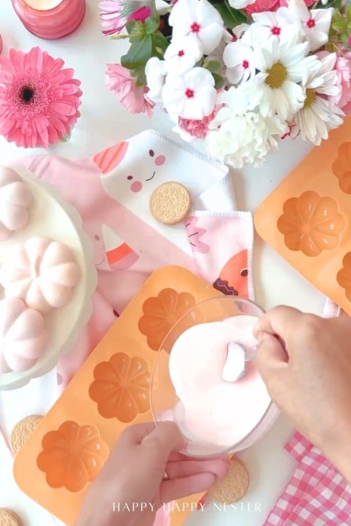 A person dips a pink marshmallow into melted coating, surrounded by flower-shaped molds, cookies, a pink floral napkin, and a bouquet—creating sweet magic for an Oreo Halloween treats recipe on a pastel-themed table.