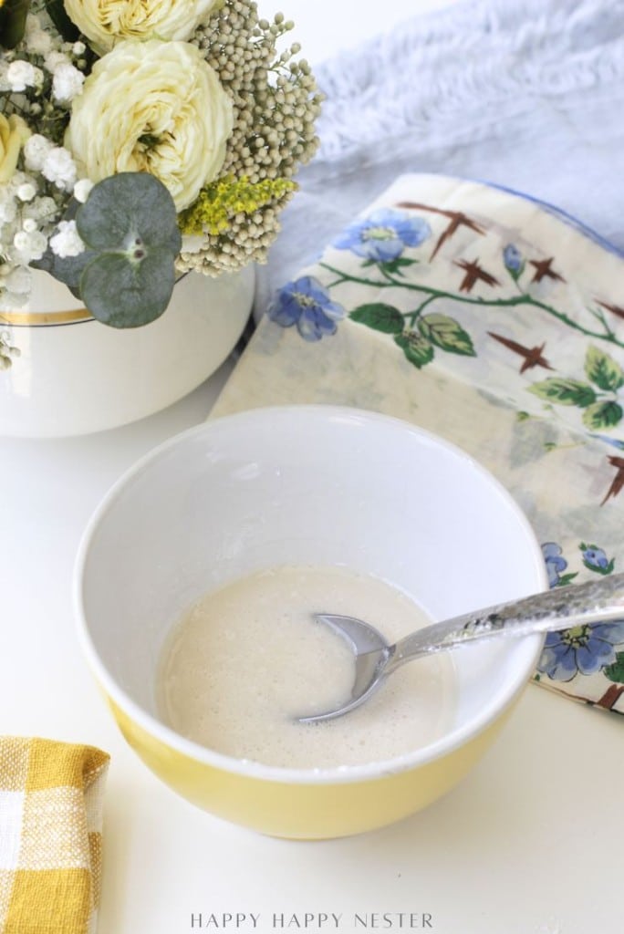 A bowl with a spoon resting inside holds a small amount of white mixture—perfect for a mini waffle donuts recipe. The bowl sits on a white surface near a yellow cloth, floral napkin, and vase of flowers.